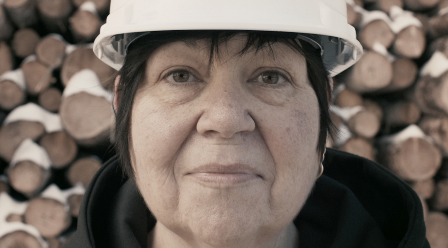 Close up of Tina Rasmussen’s face, Chief Business Officer of Meadow Lake Tribal Council, in a white hard hat with stacked lumber behind her. 