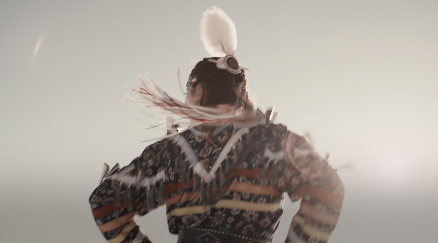 An Indigenous woman wearing a jingle dress and feathers in her hair dances outdoors, with her back to the camera.