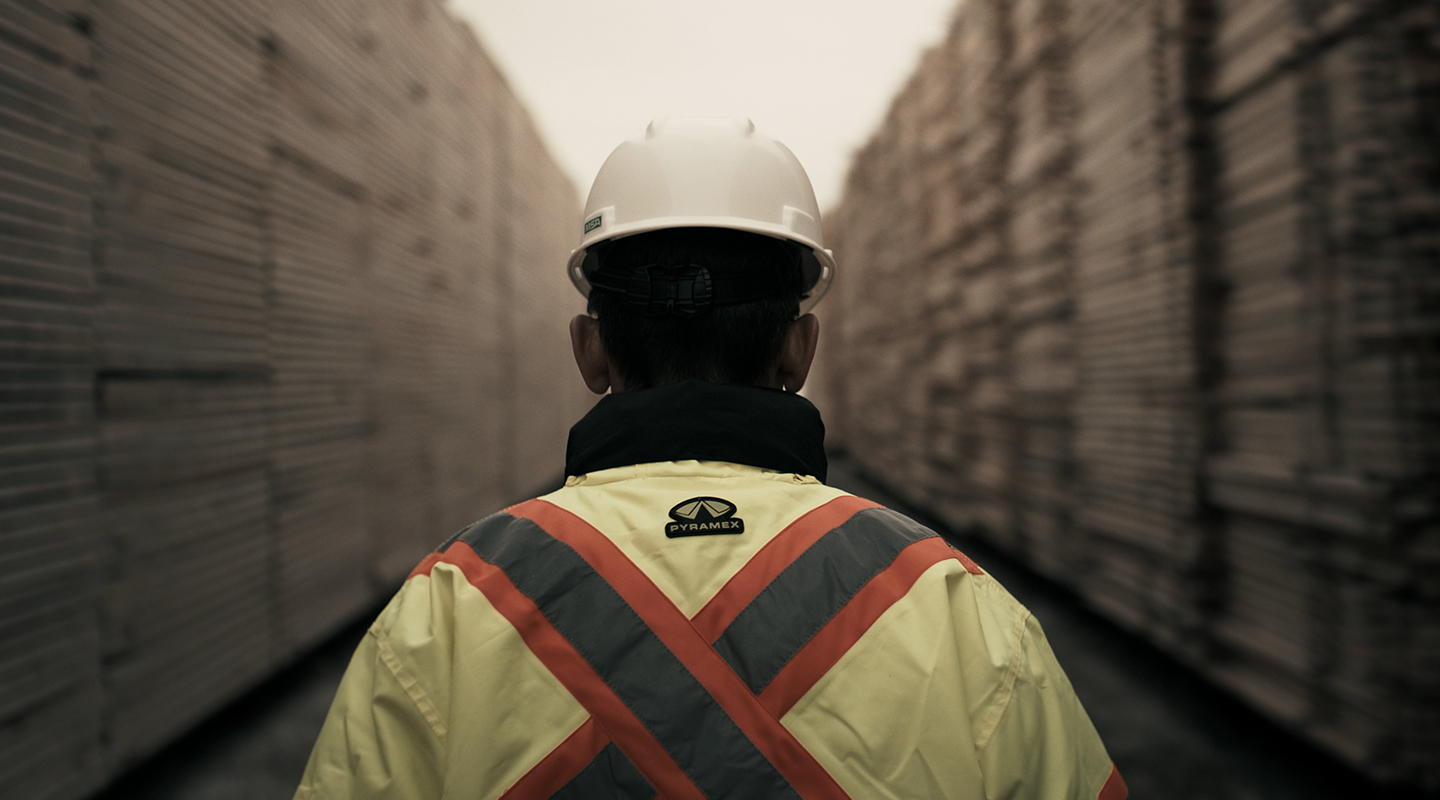 A man wearing a hard hat and security vest standing in between two large stacks of lumber, with his back to the camera.