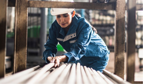 A woman wearing coveralls and a safety helmet bending down to move steel poles from a shelf.